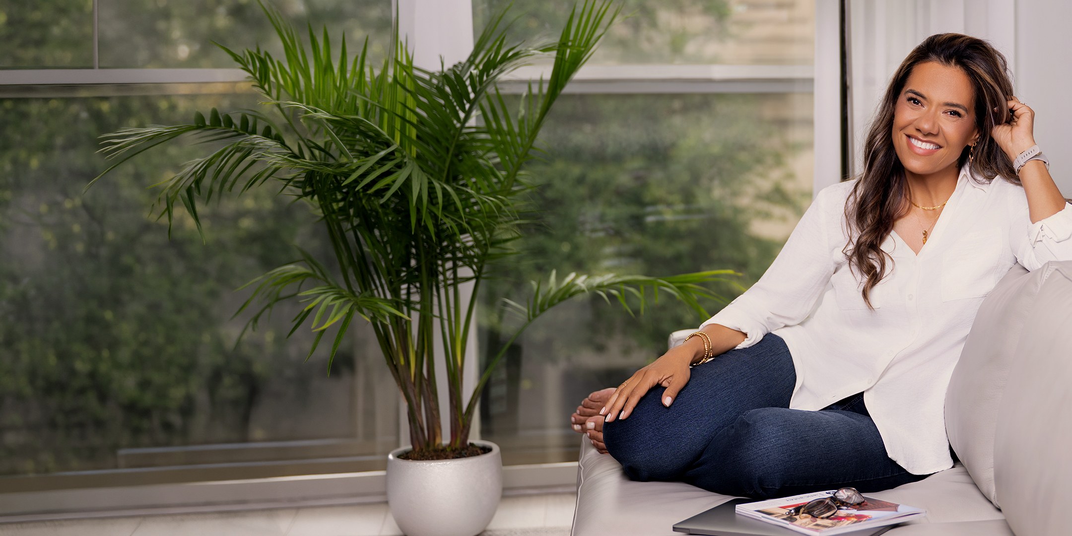 Christiane Drew smiling, seated on a sofa beside a palm plant in a bright, sunlit room